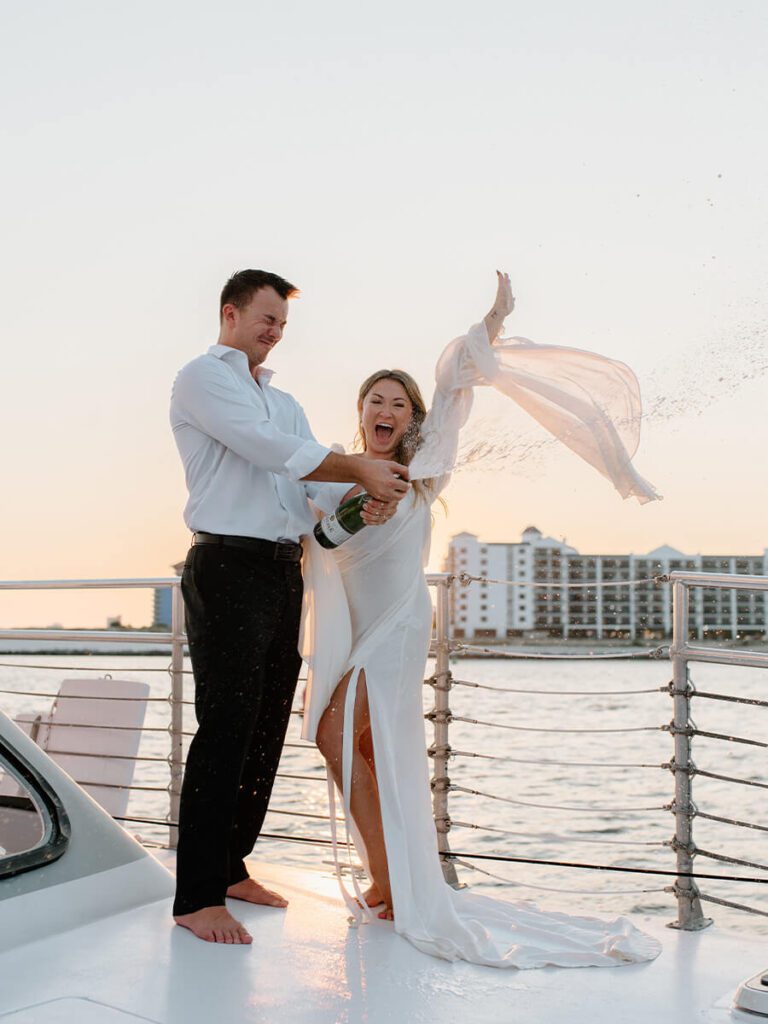 Couple popping a bottle of champagne on a sailboat in wedding attire